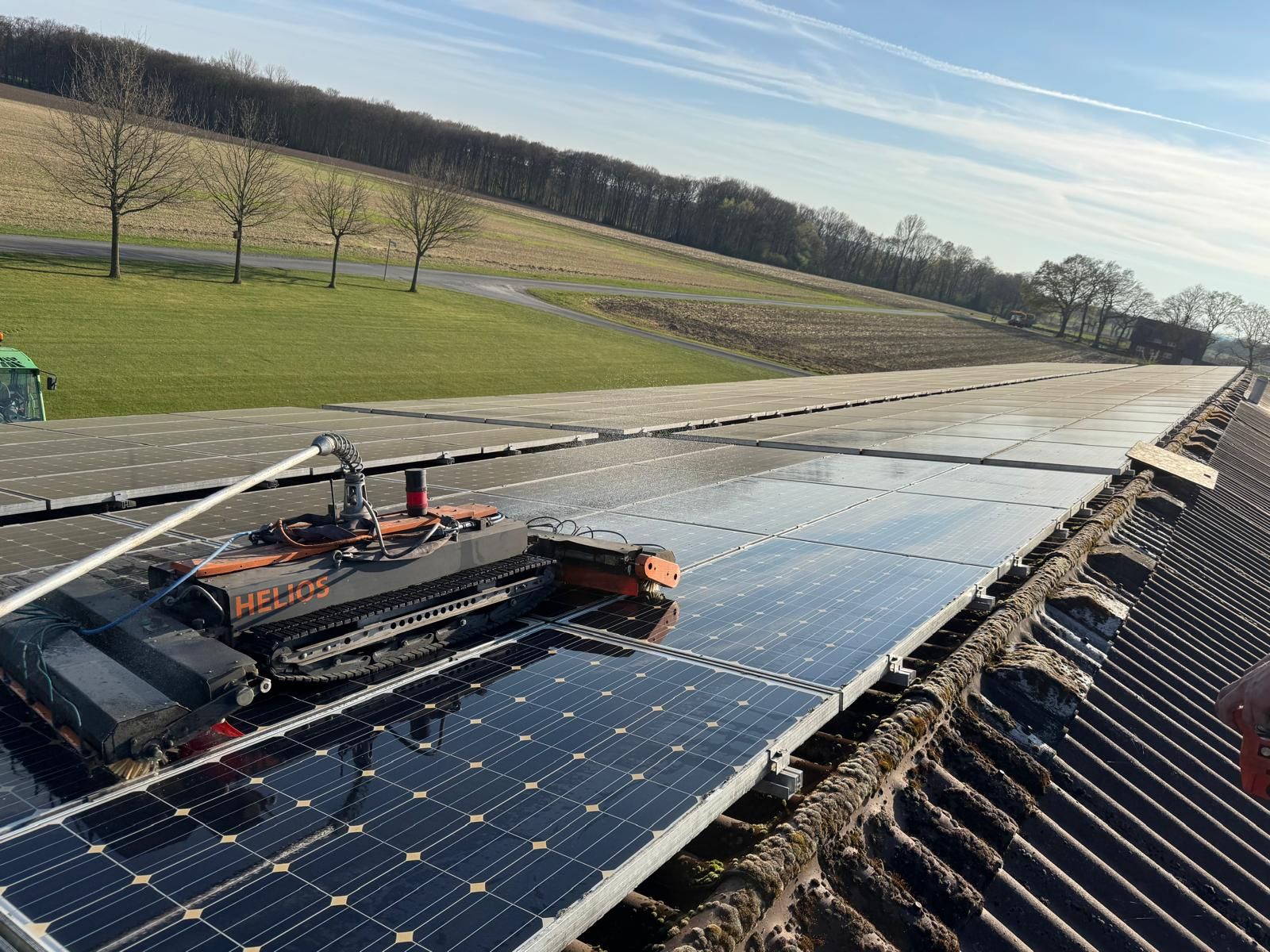 Reinigungsroboter auf einer langen Reihe von PV-Modulen auf einem Dach, dahinter Felder und eine Baumreihe im Abendlicht.