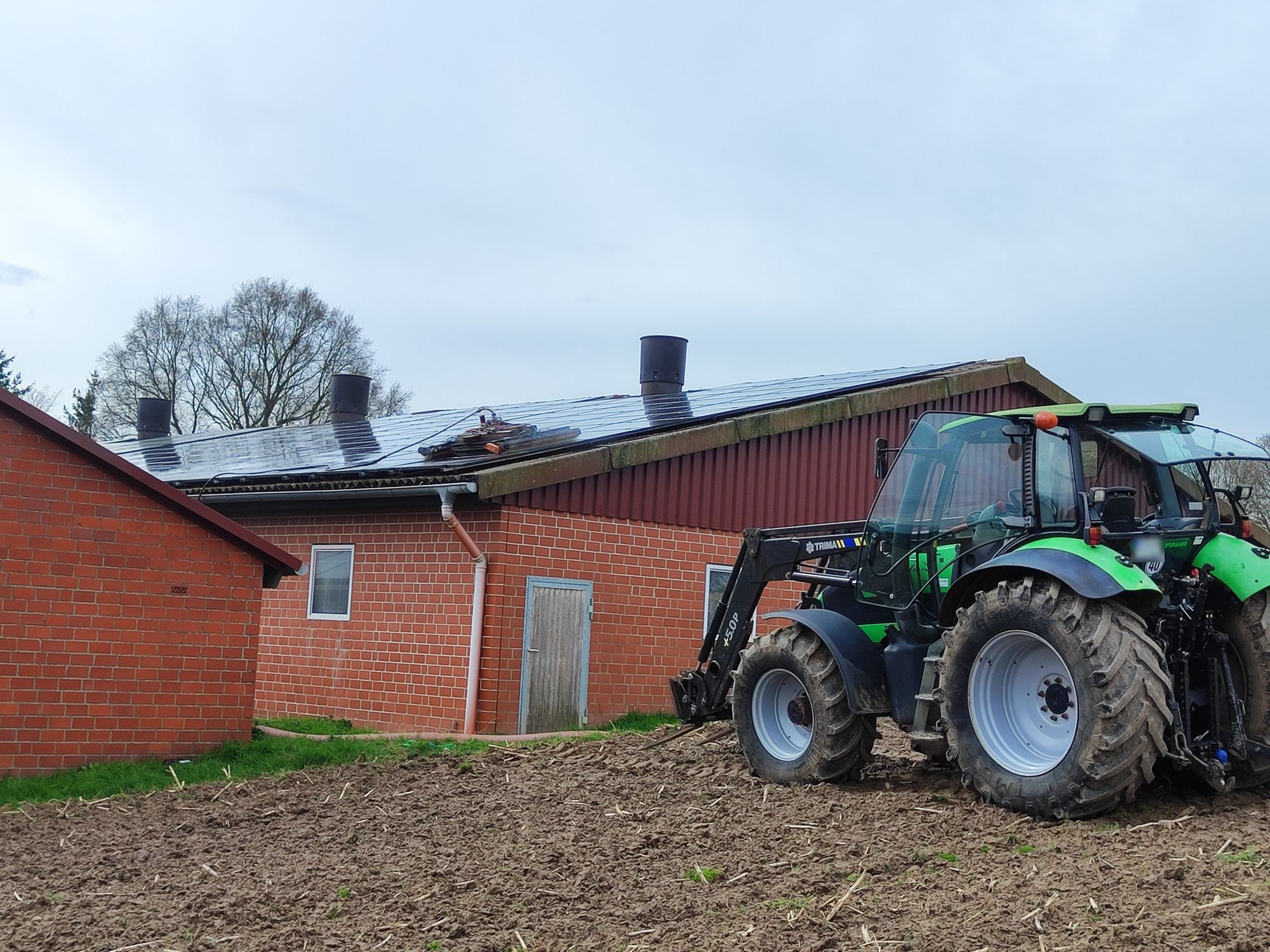 Stall aus Klinker mit Photovoltaikanlage und Abluftkaminen auf dem Dach, oben der Reinigungsroboter. Im Vordergrund steht ein grüner Traktor auf dem Acker.